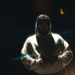 A man wearing a hoodie stands in dramatic lighting at night, hands in pockets, conveying a mysterious mood.