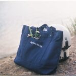 A blue tote bag with wildflowers and a helmet by the tranquil riverbank.