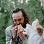 A man happily eats a sandwich outside in a green and natural setting.